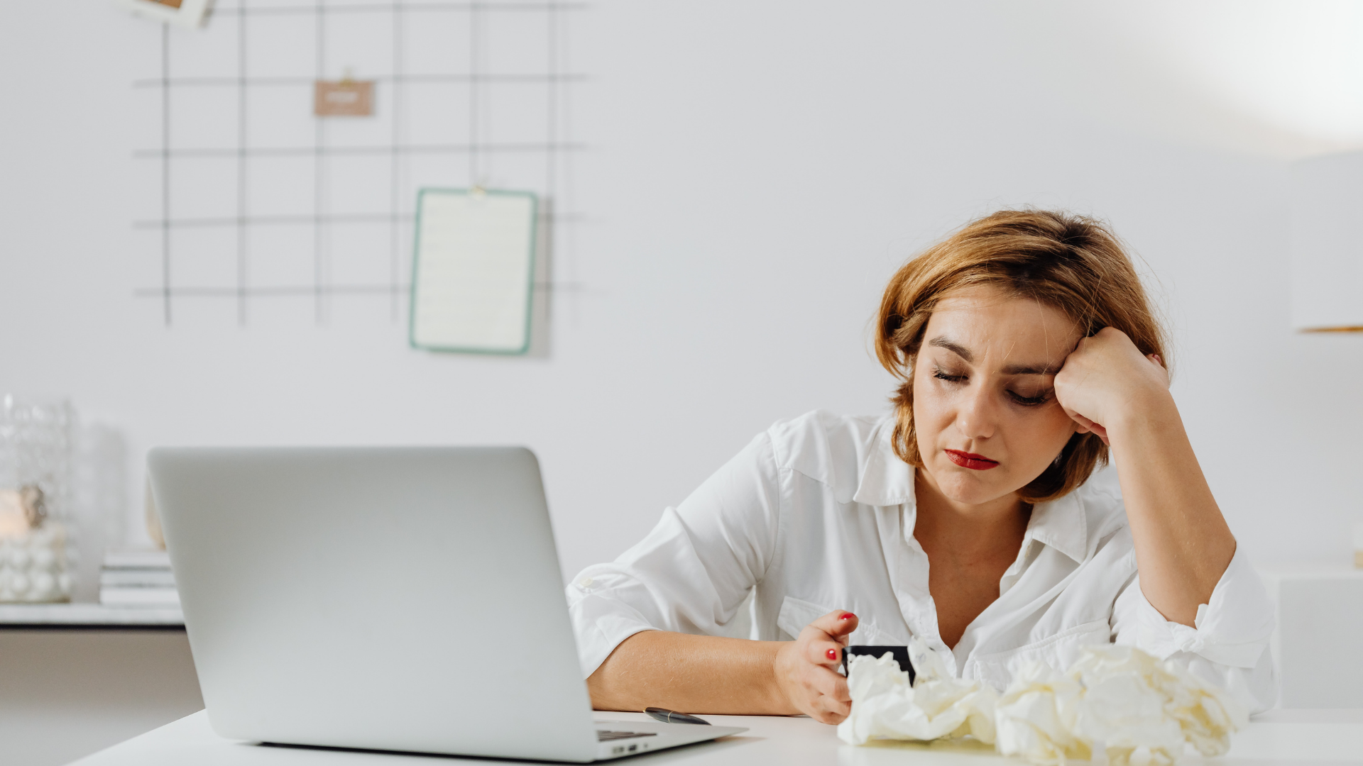 Woman with laptop looking at phone, resting head on hand. White shirt, tissue, modern office.