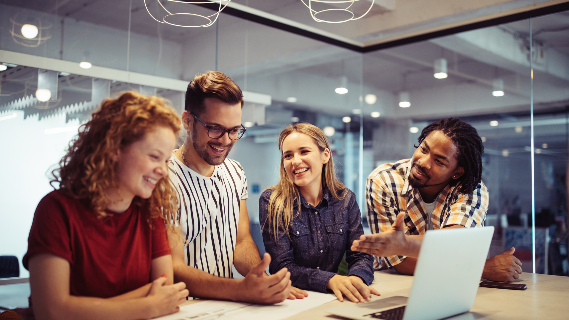 Four diverse people collaborating at a table with a laptop, smiling, in a modern office.