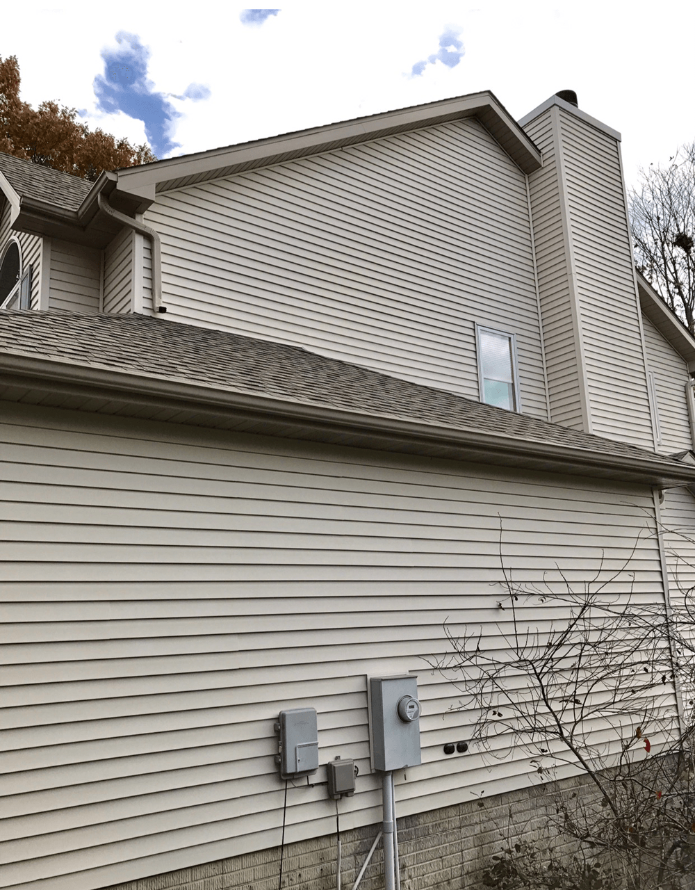 Tan siding of a two-story house with a chimney; overcast sky and sparse foliage visible.