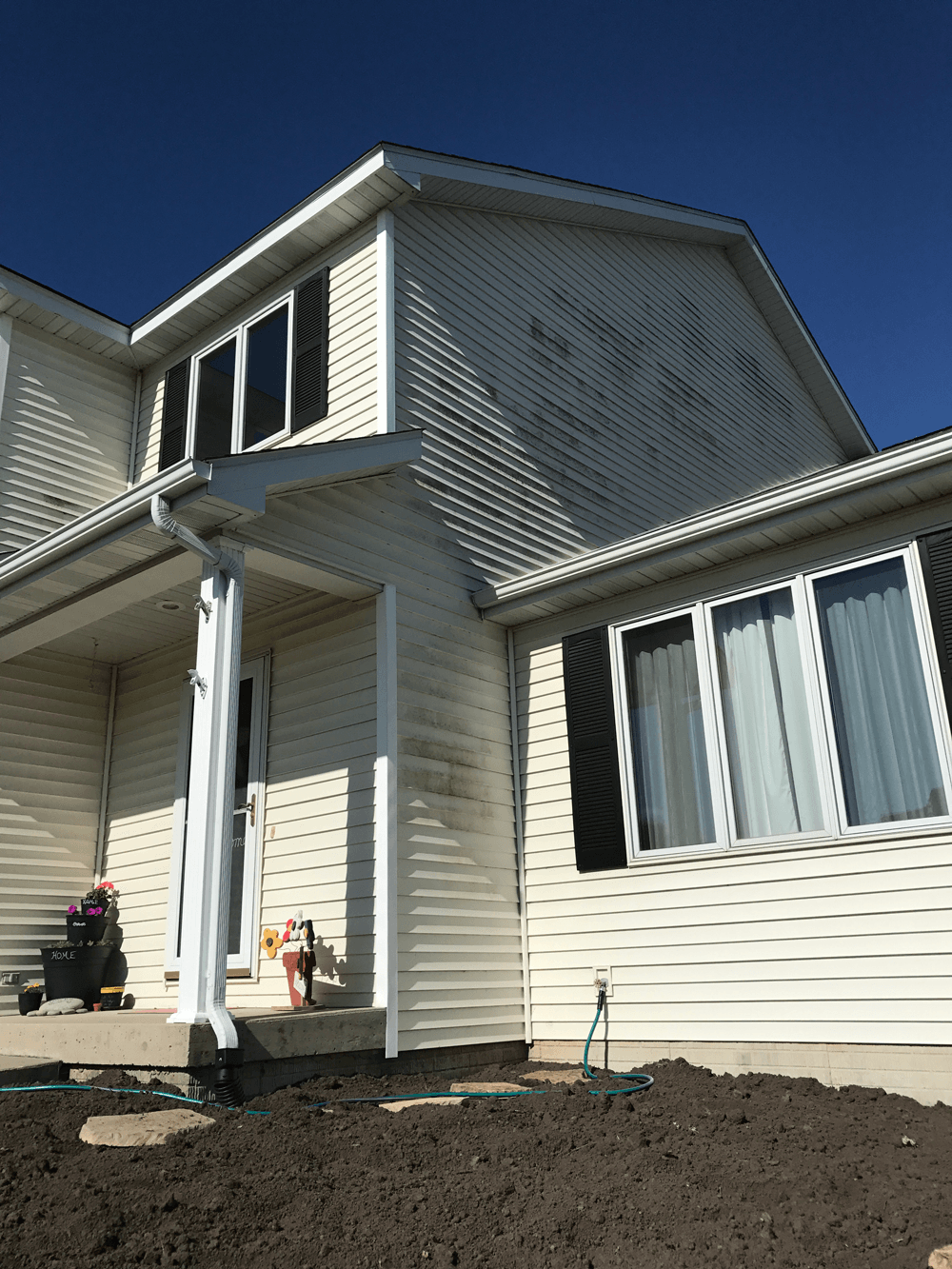 Two-story house with white siding; dirty areas; front porch with a column; dark soil in the yard; clear blue sky.