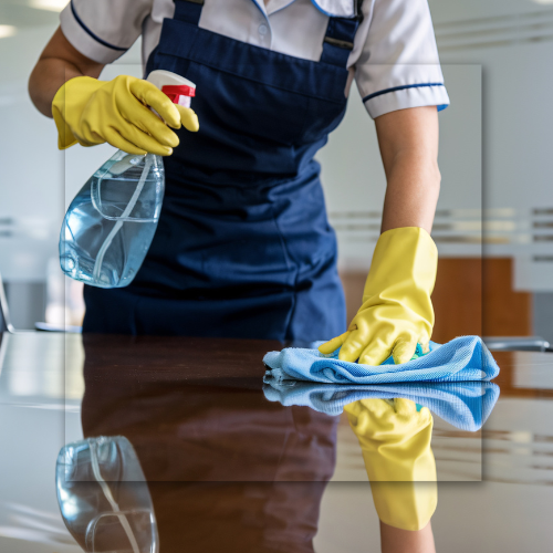 A person wearing yellow gloves and an apron is cleaning a desk with spray and a blue cloth.