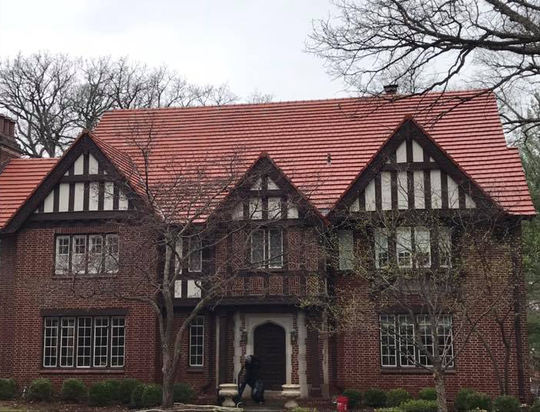 Brick Tudor-style house with red tile roof and white trim. Two people stand at the front door.