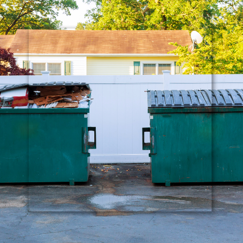 Two green dumpsters, one overflowing, behind a white fence, with a house in the background.
