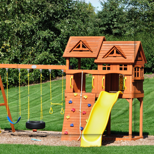 Wooden playset with swings, rock climbing wall, slide, and clubhouse in a grassy yard.