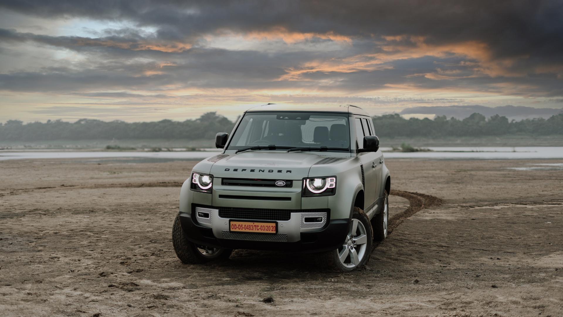 Green Land Rover Defender on a dirt field with a cloudy sky in the background.