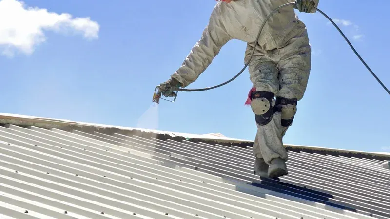 Person in protective suit spraying a white coating onto a metal roof under a blue sky.