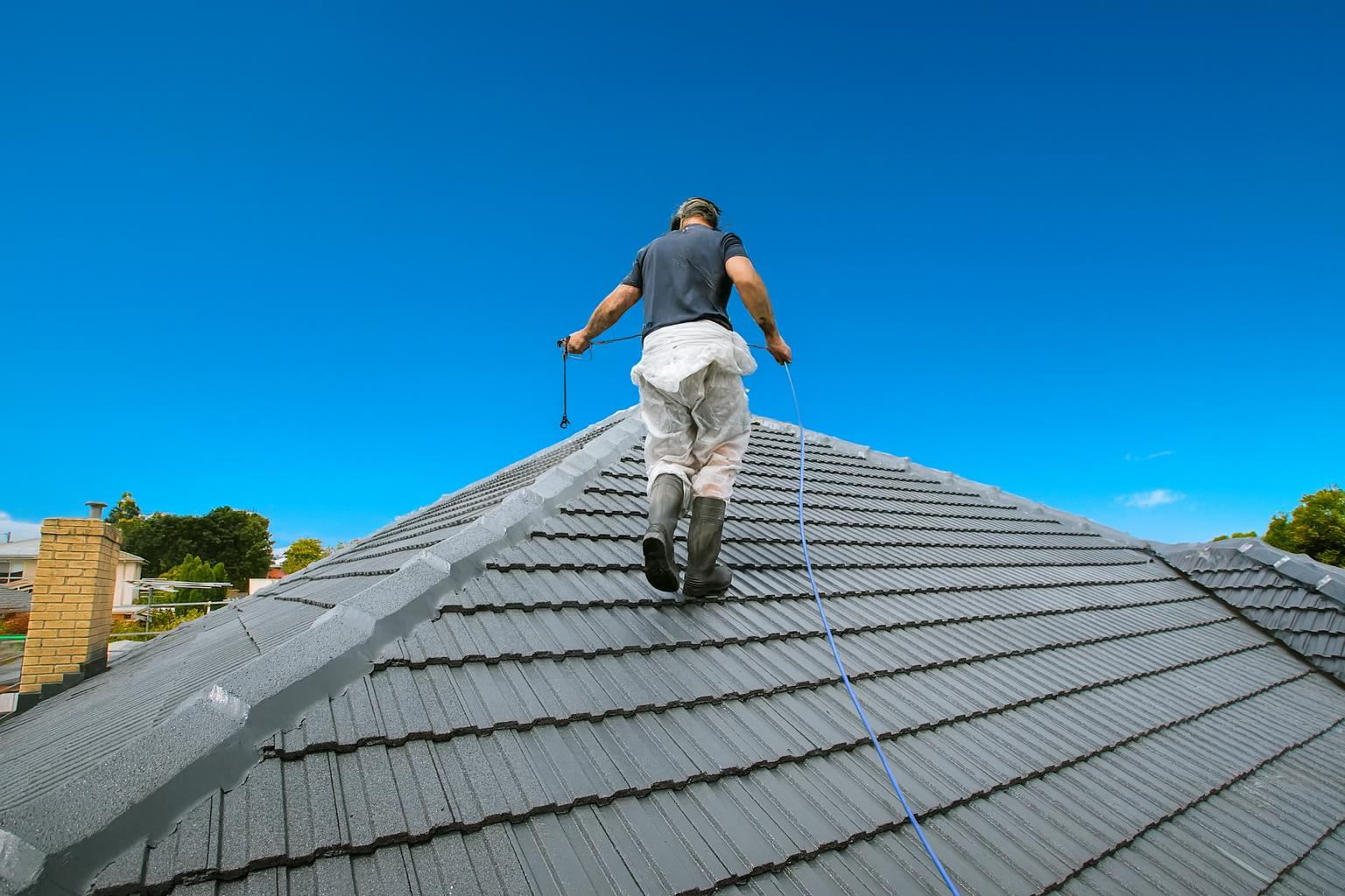 Roofer in work clothes walks on a gray tiled roof under a bright blue sky.