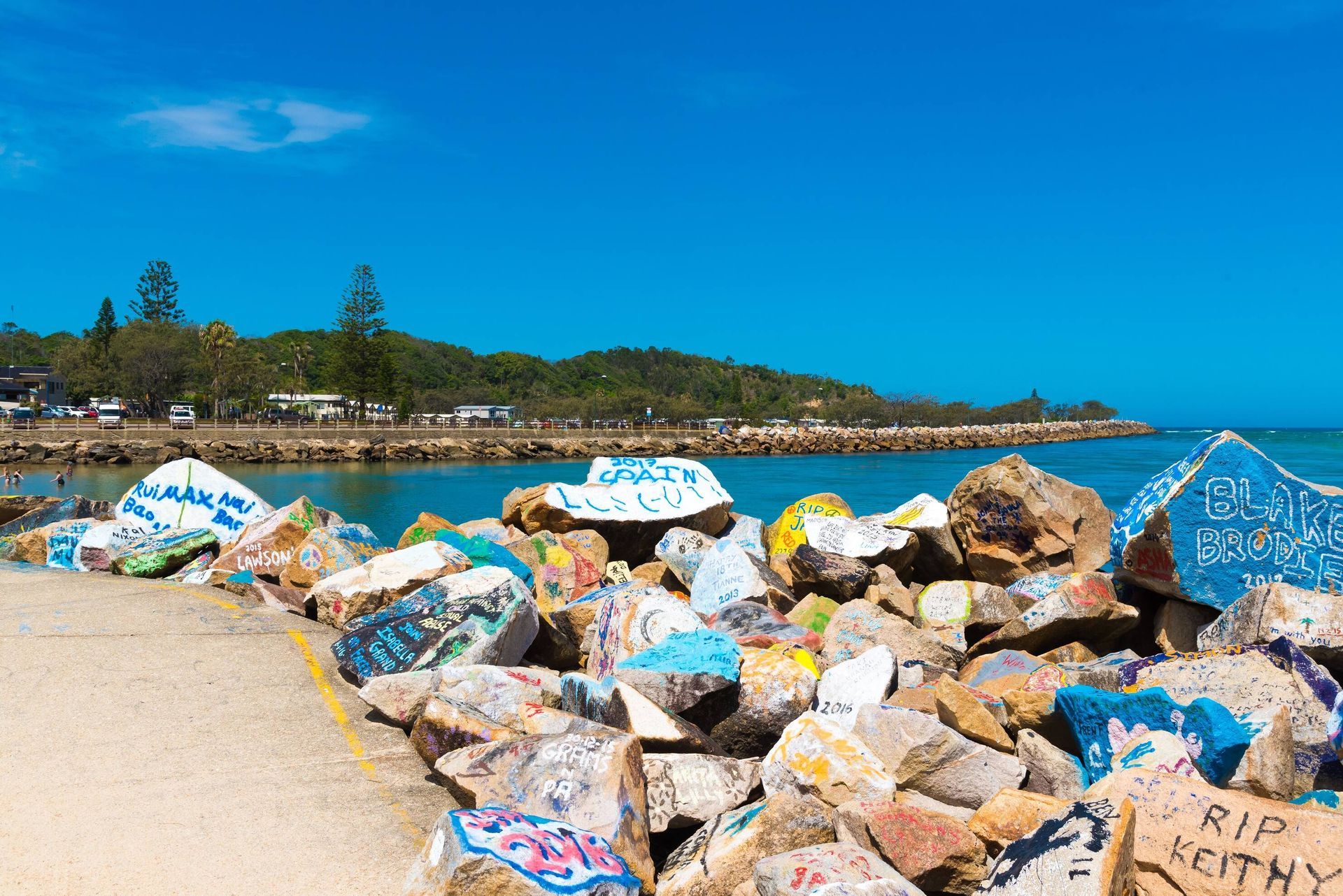 Nambucca Head - Colorful graffiti-covered rocks line a coastal edge under a bright blue sky, with trees and buildings in the distance.