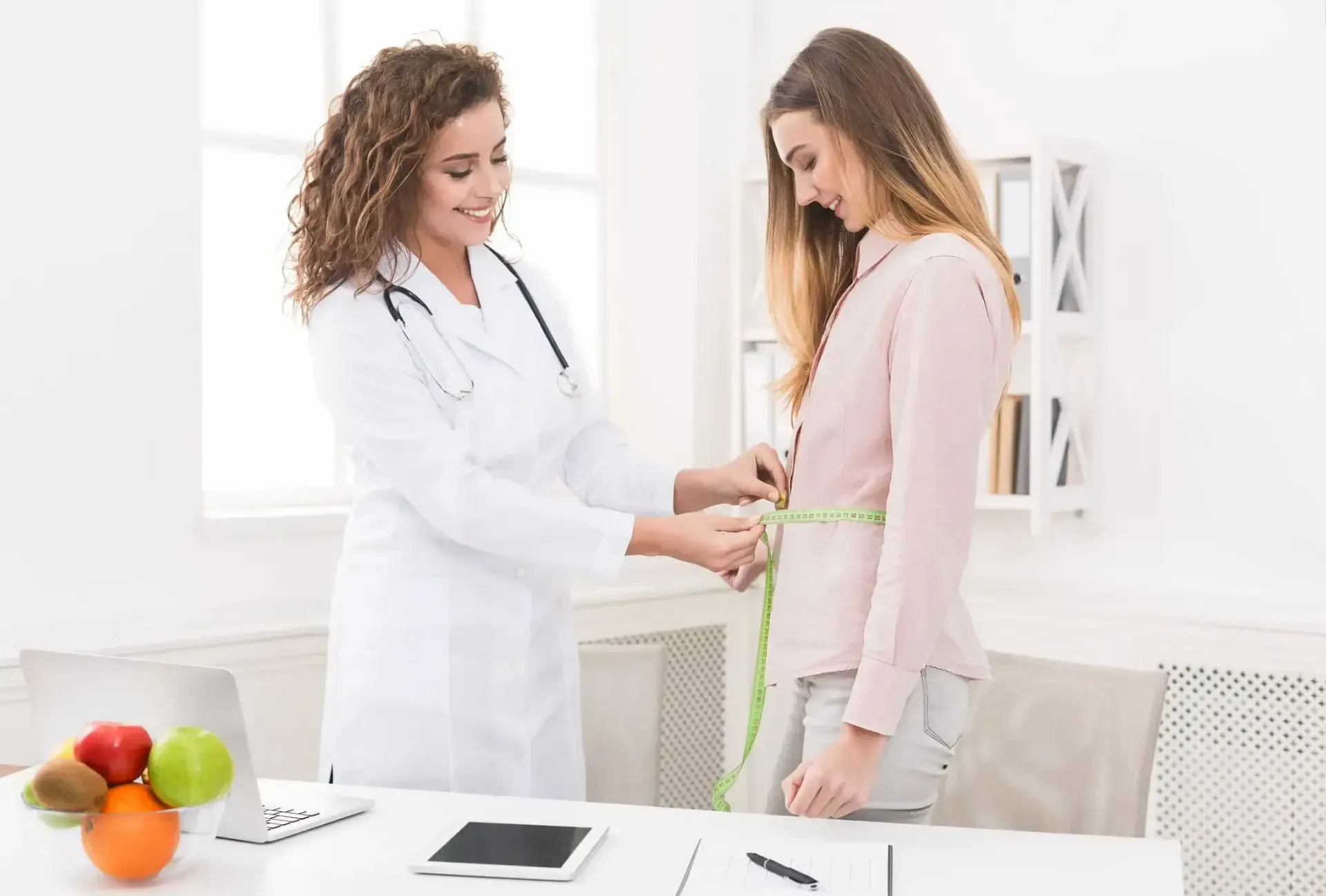 A doctor measures a patient's waist with a tape measure in a bright office.