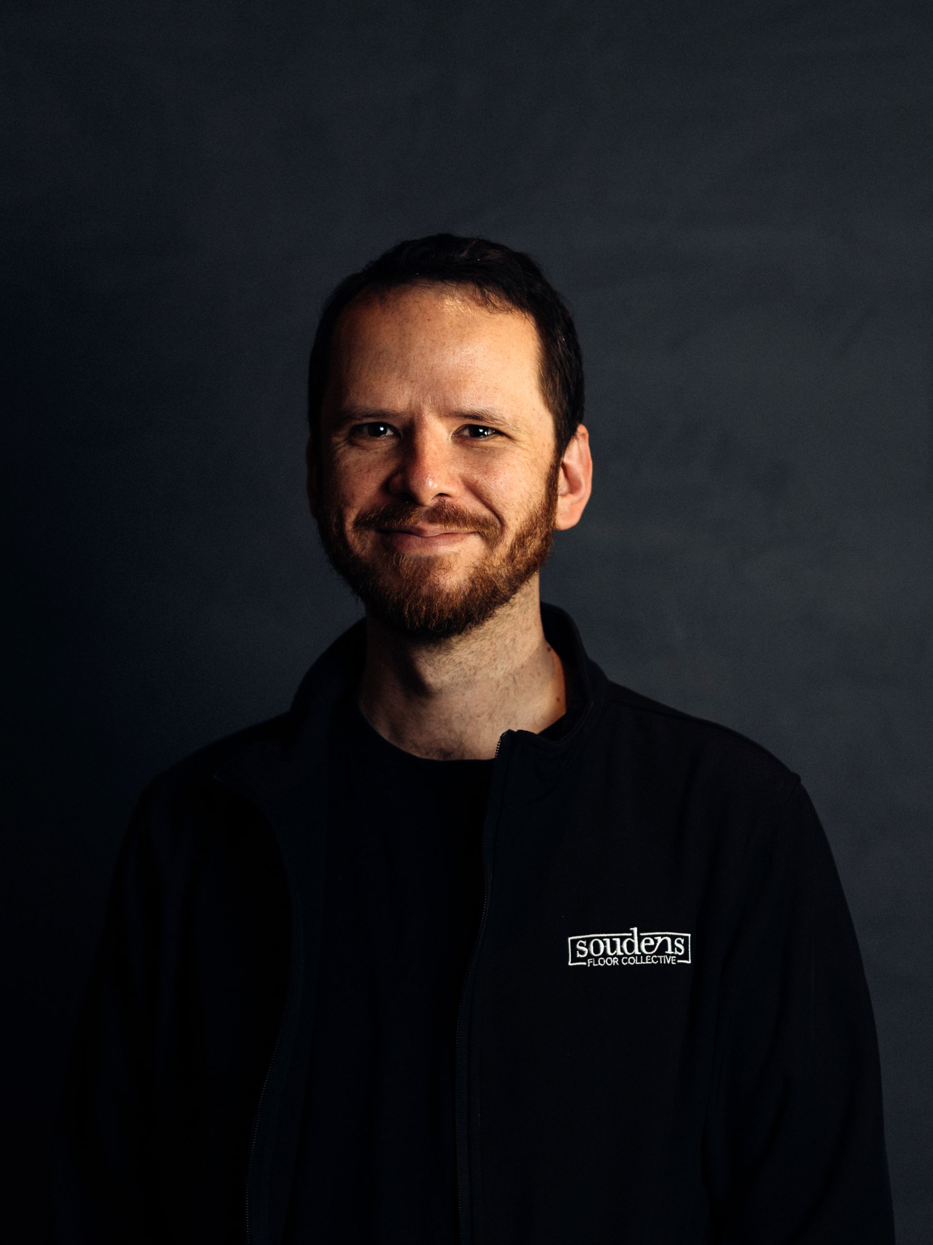 A man with a beard is wearing a black jacket and smiling.