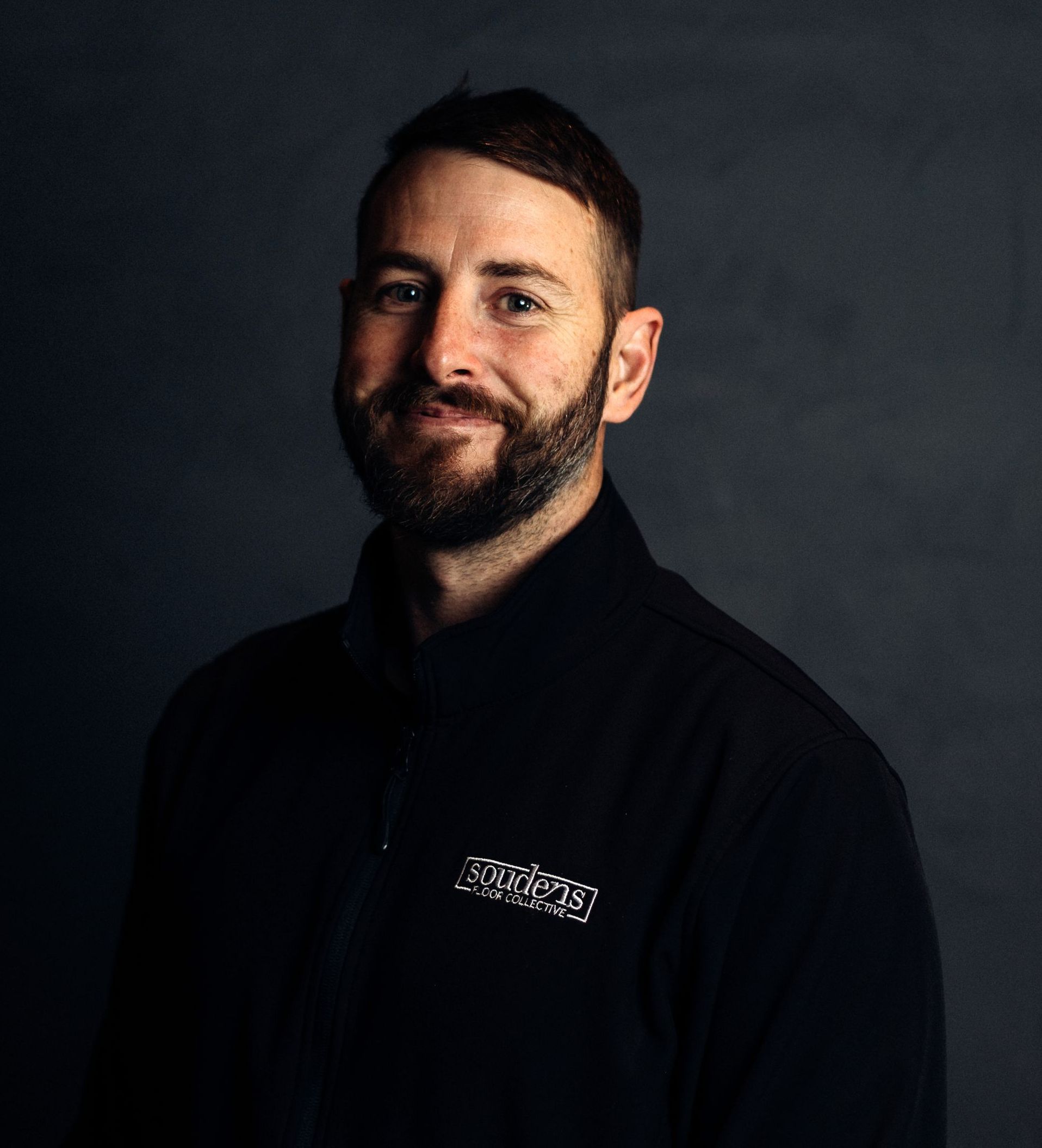 A man with a beard is wearing a black jacket and smiling for the camera.
