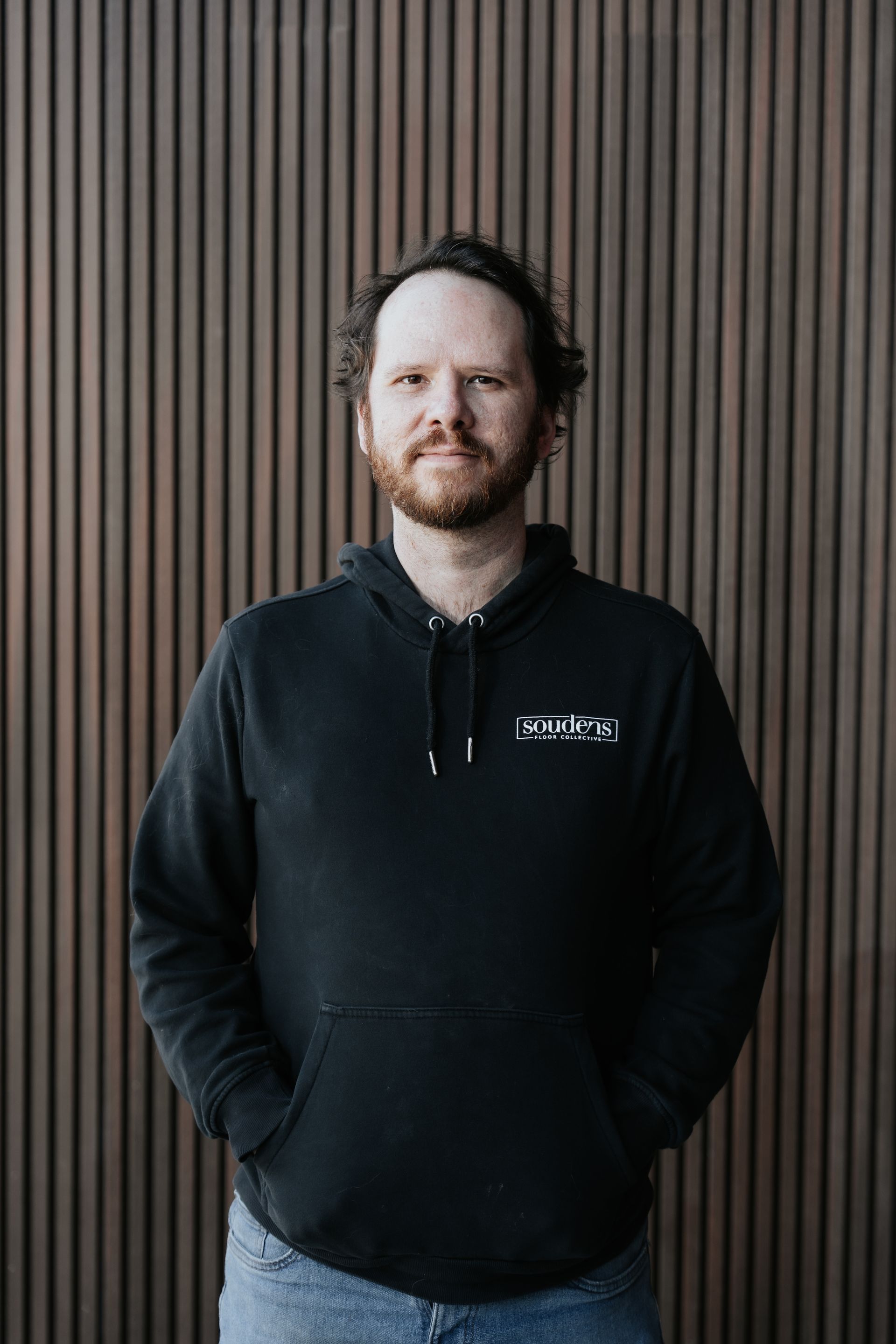 A man with a beard is wearing a black jacket and smiling.