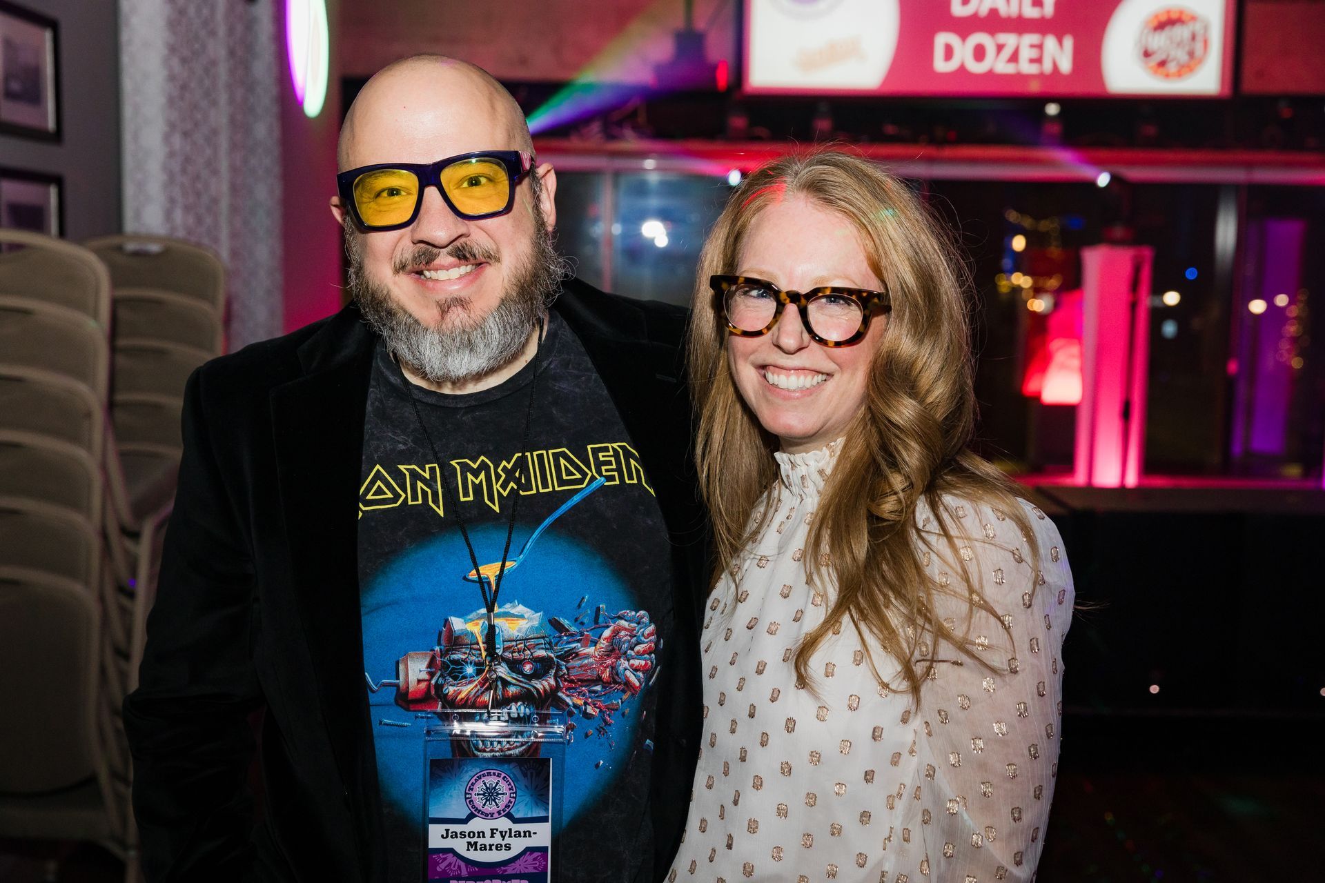 Bald man in blue shirt and yellow glasses, gestures with hands, against black background.