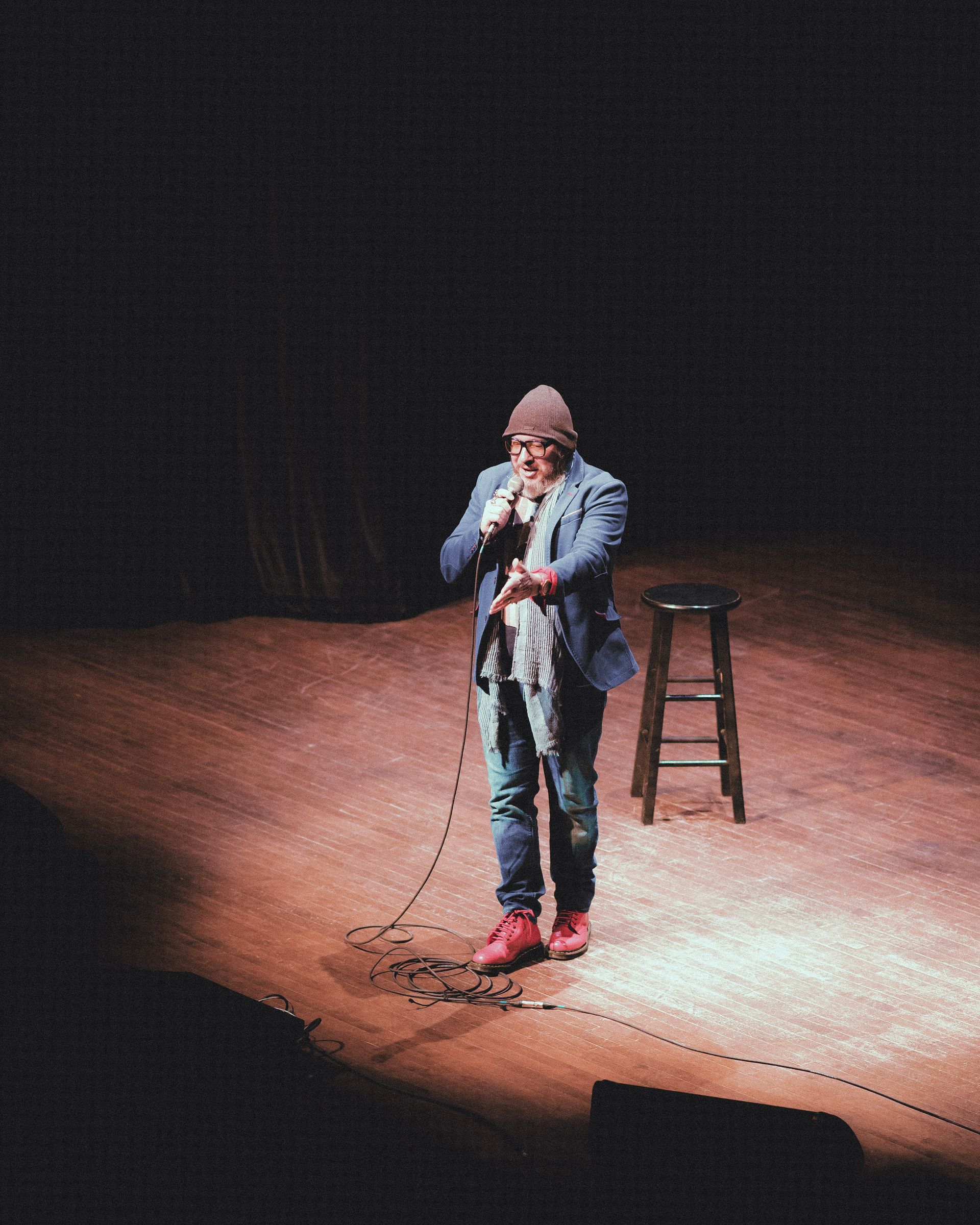Comedian on stage, holding mic, in spotlight, stool to his left, dark background.