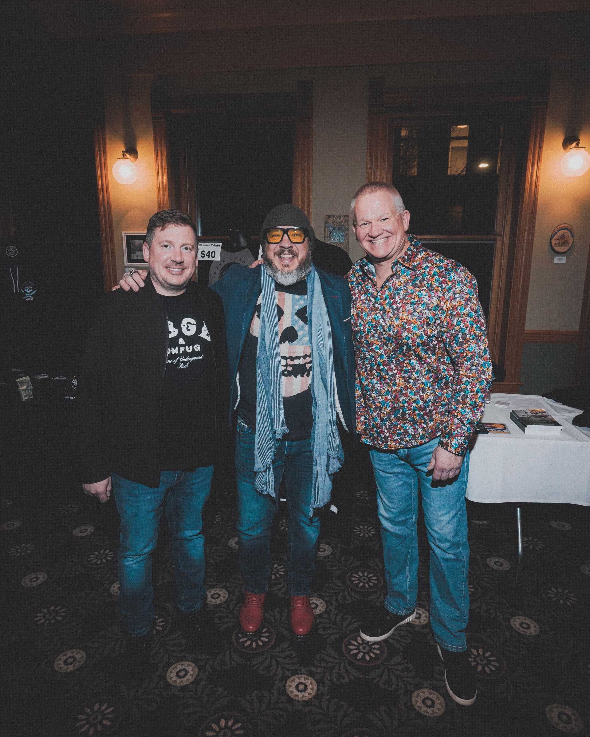 Three men posing for a photo indoors. One in a floral shirt, one in a hat, and one with arm around the other.