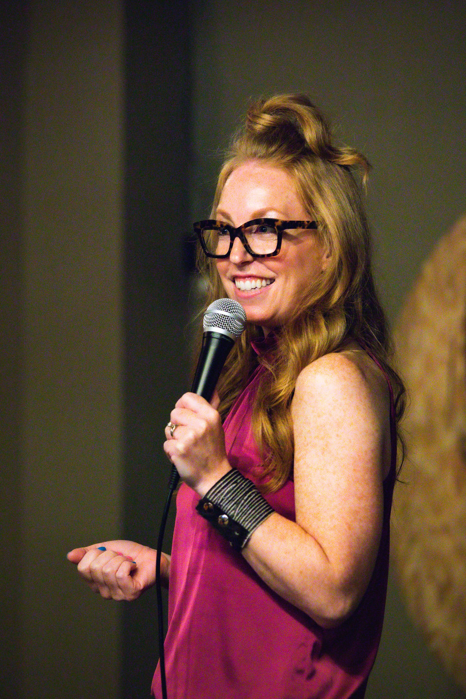 Woman with glasses, holding a microphone, smiling on stage, wearing a pink top.