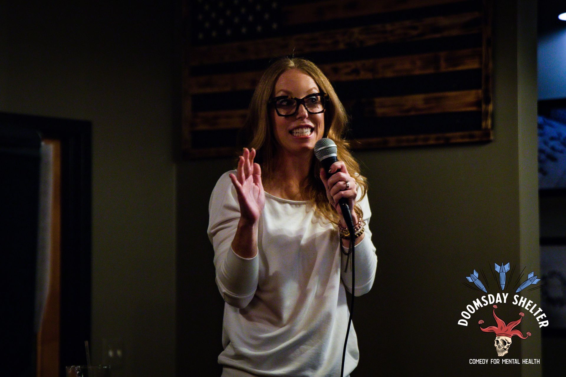 Woman with glasses, holding a microphone, gesturing while speaking at a venue with a US flag background.