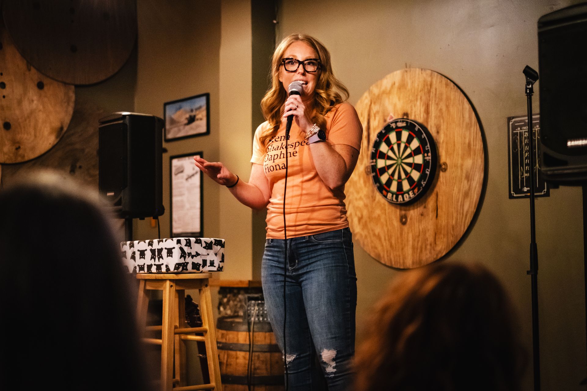 Woman with red hair and glasses performing stand-up comedy in a bar, holding a microphone.