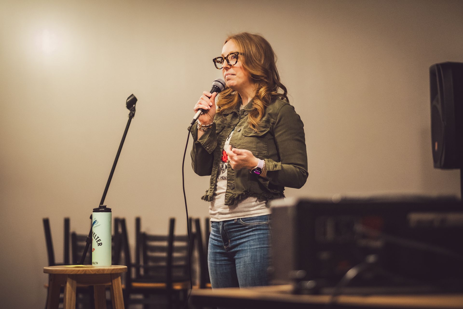 Woman with glasses speaks into a microphone on stage; olive green jacket, jeans, neutral background.
