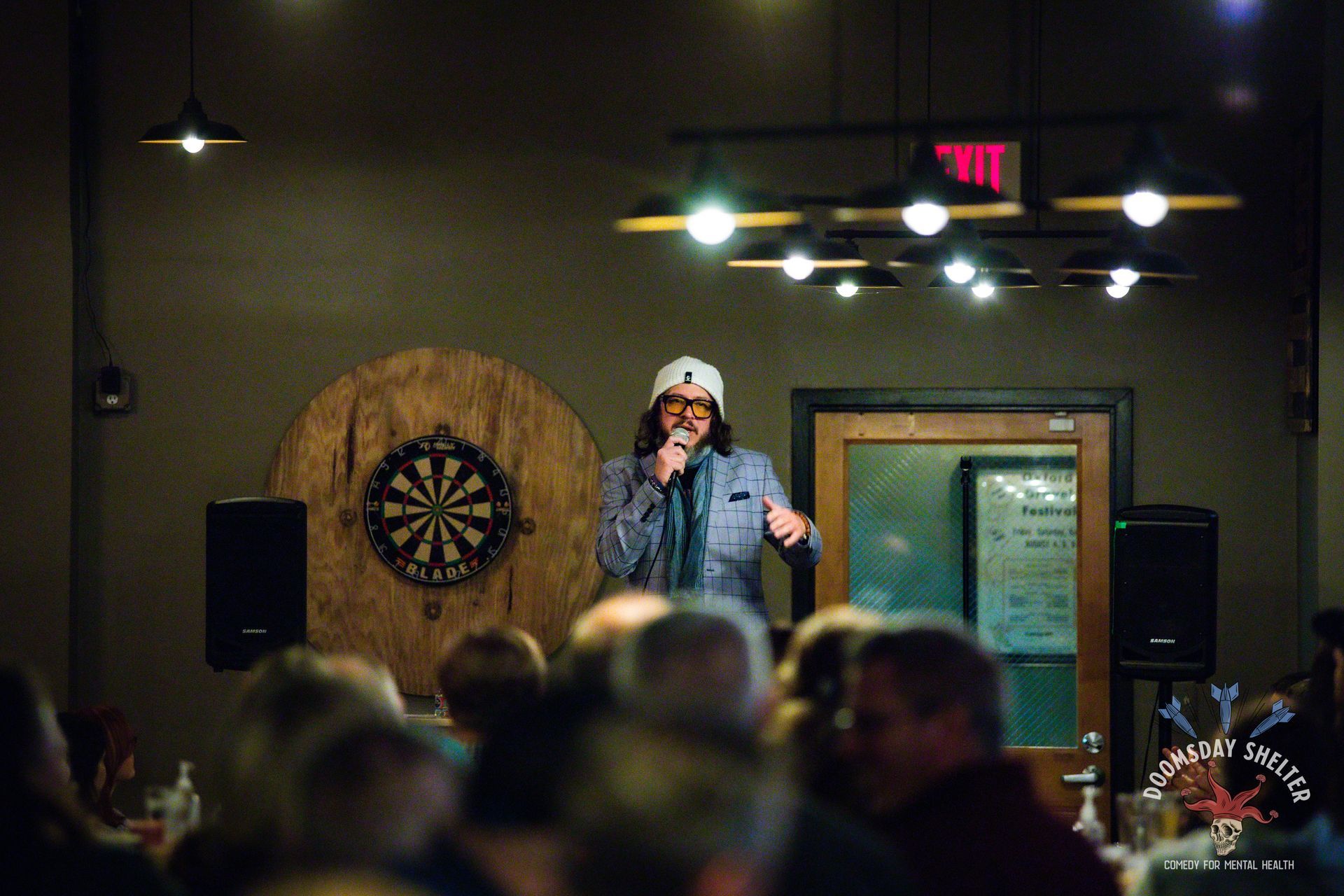 Comedian on stage, wearing a hat, jacket, and glasses, in front of a dartboard. Crowd in the foreground.