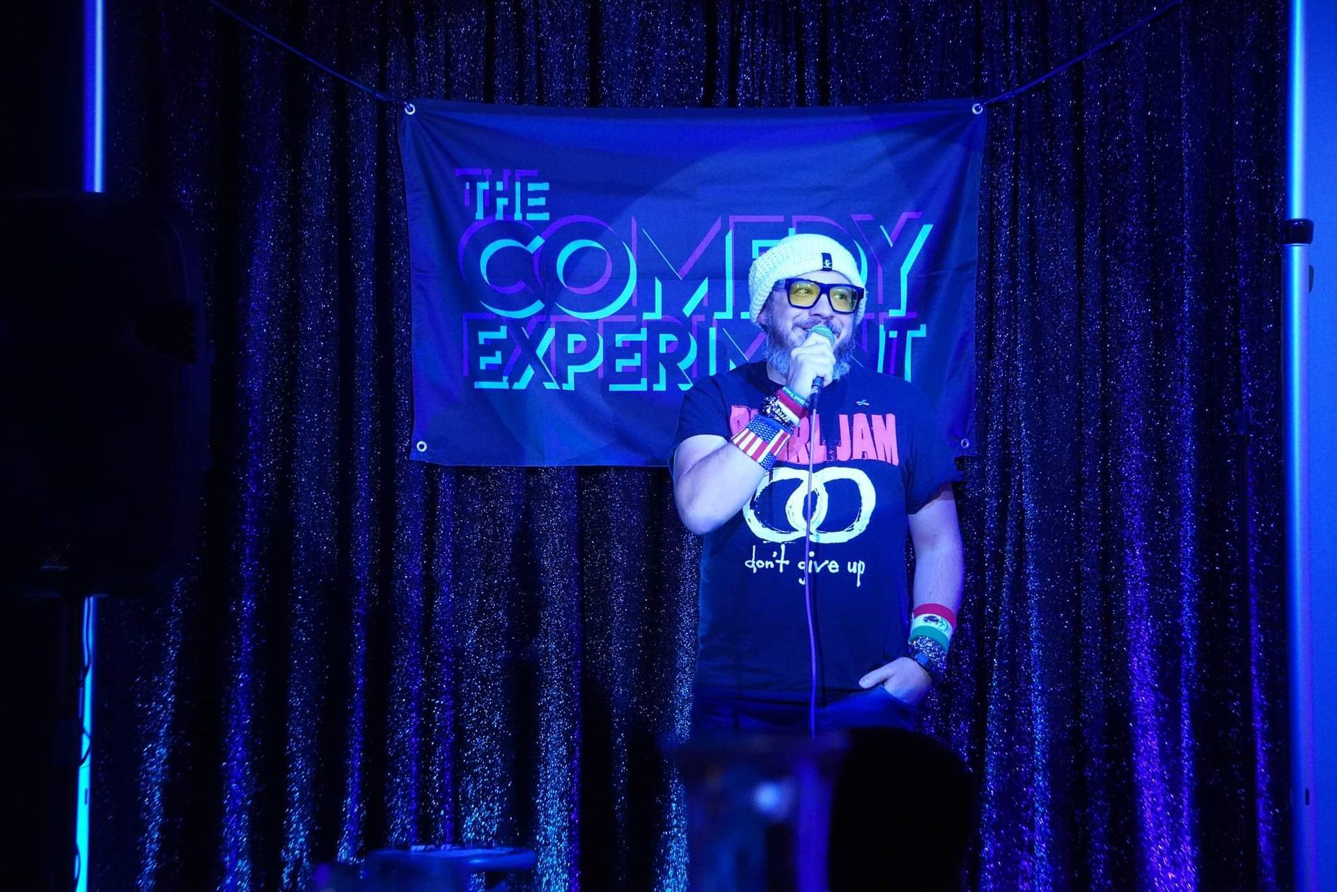 Comedian on stage, wearing sunglasses, microphone in hand, in front of a 