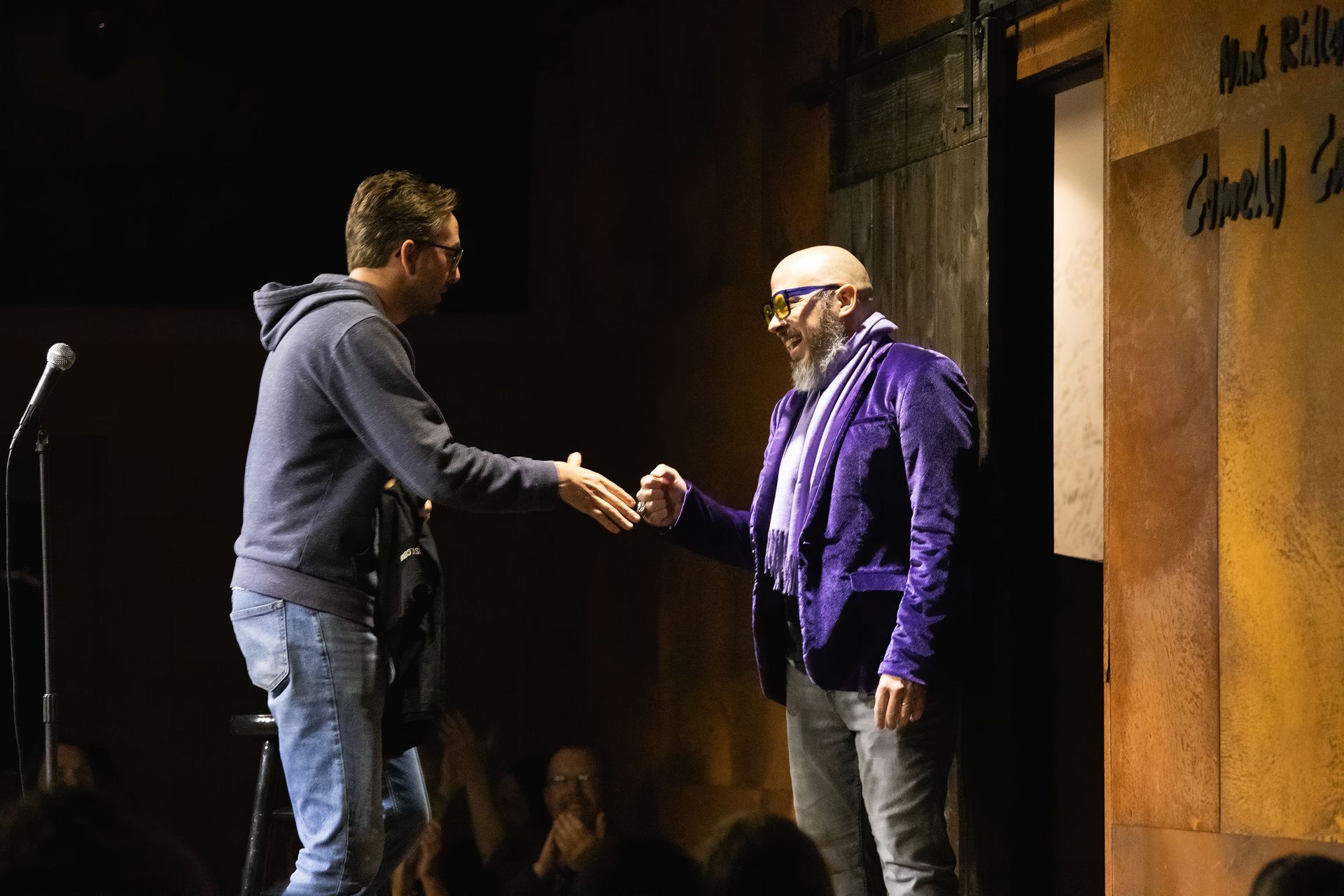 Two men shaking hands on stage. One in a blue hoodie, the other in a purple jacket. Dimly lit comedy club.