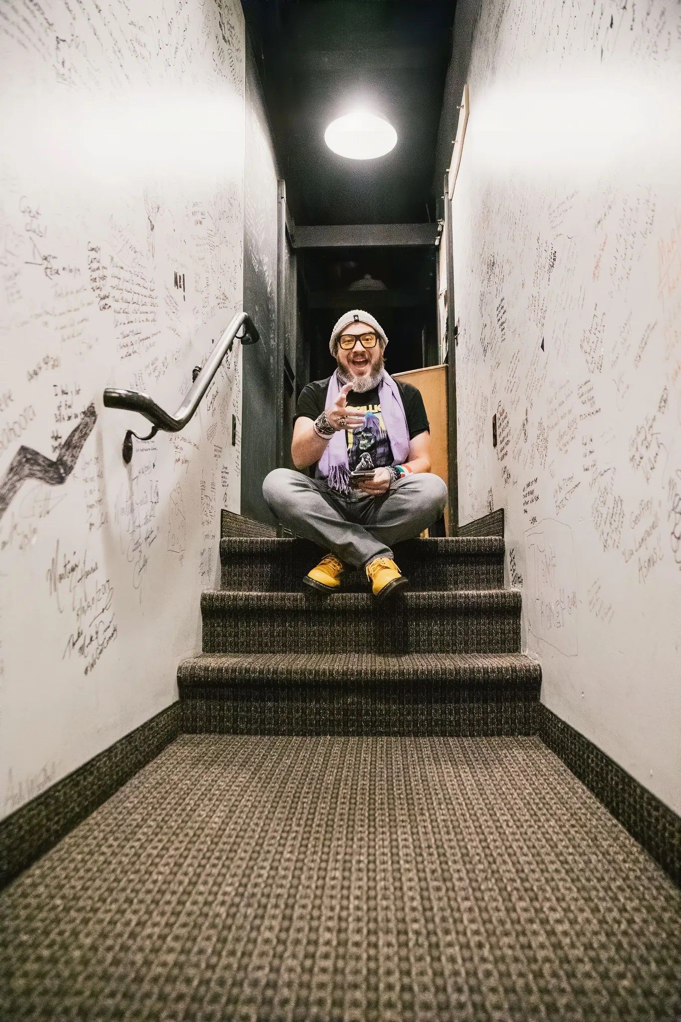 Person sitting on stairs, smiling, holding a drink. They wear yellow shoes and a purple scarf, stairs in a narrow hallway.