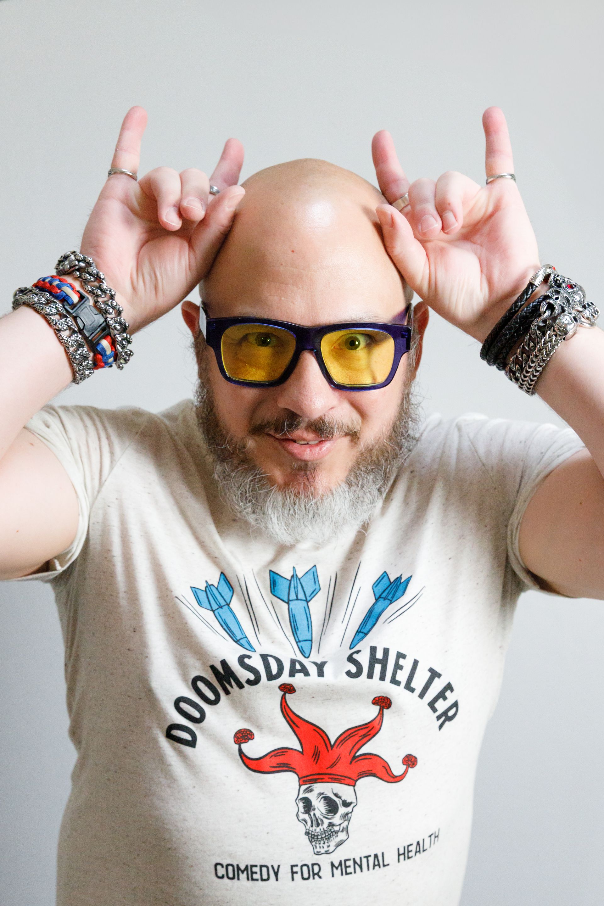 Bald man with beard, yellow glasses, and bracelets makes a rock-on gesture. Wears