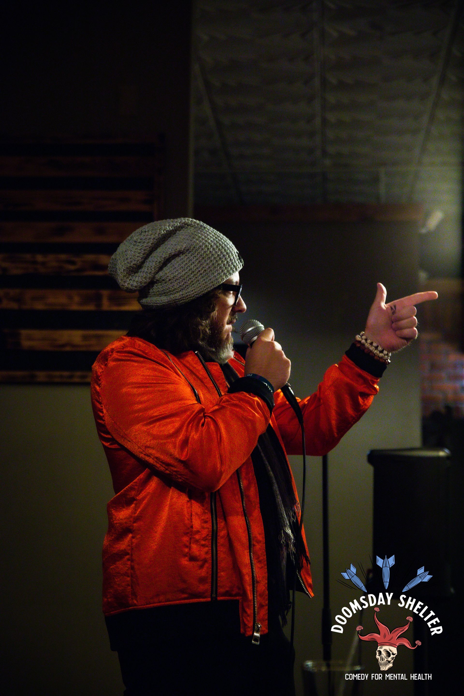 Person with a microphone in a red jacket and beanie points to the side. Indoor setting.