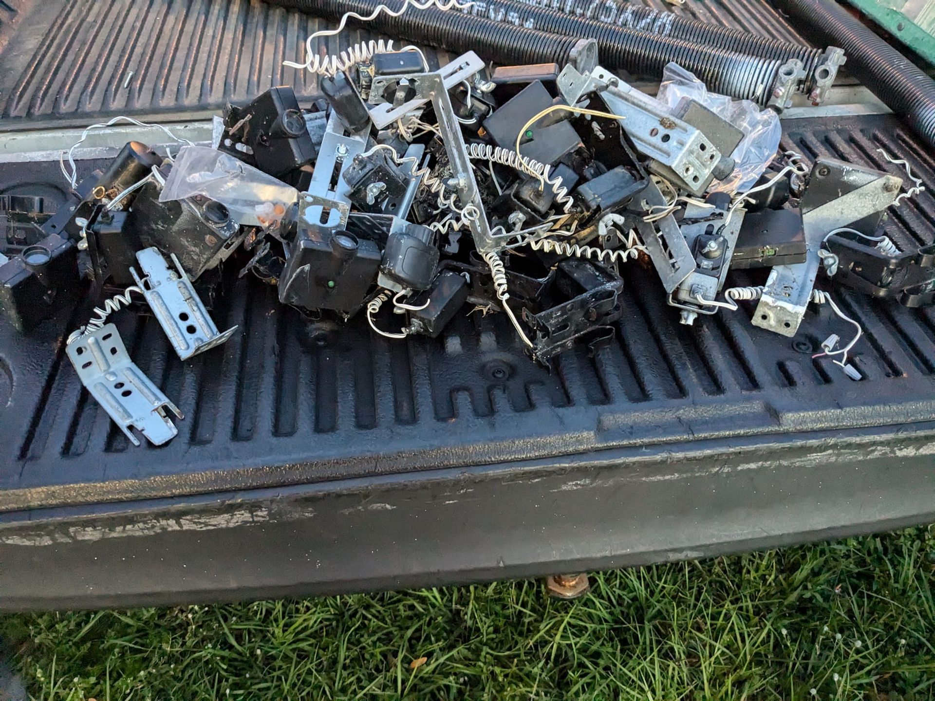 Pile of black electrical components and metal parts in a truck bed, against a backdrop of green grass. Pile of black electrical components and metal parts in a truck bed, against a backdrop of green grass.