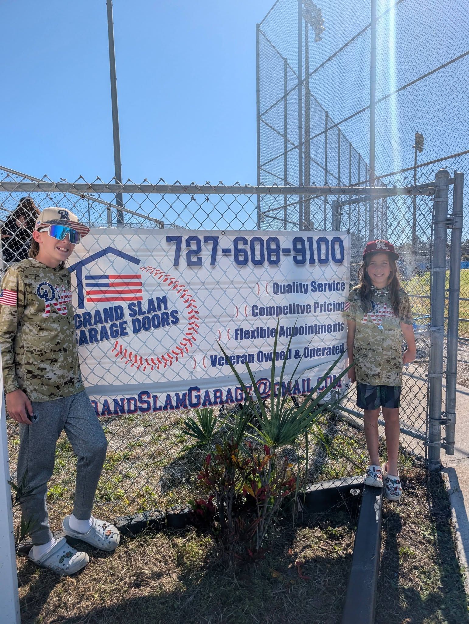 Two children stand near a chain link fence with a Grand Slam Garage Doors sign. They wear matching camouflage shirts.