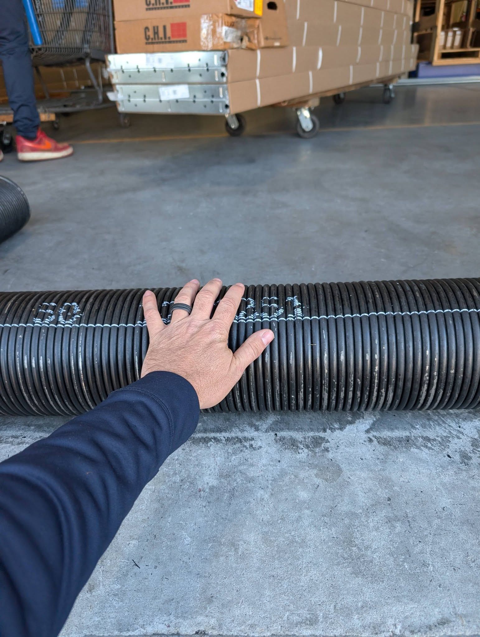 Hand on a large, black, corrugated hose on a concrete floor in a warehouse setting.
