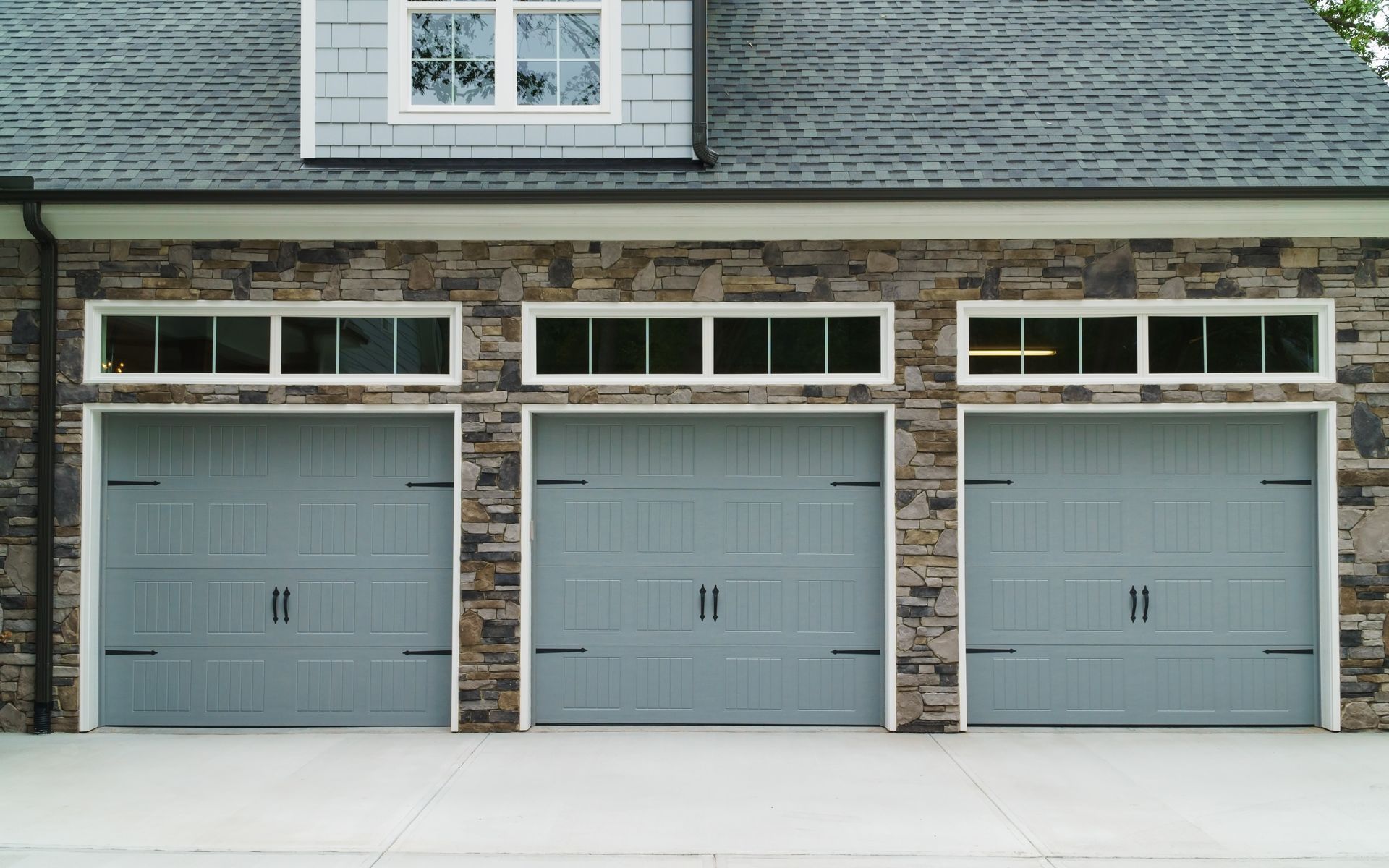 Three gray garage doors with rectangular windows above, set in a stone facade. Three gray garage doors with rectangular windows above, set in a stone facade.