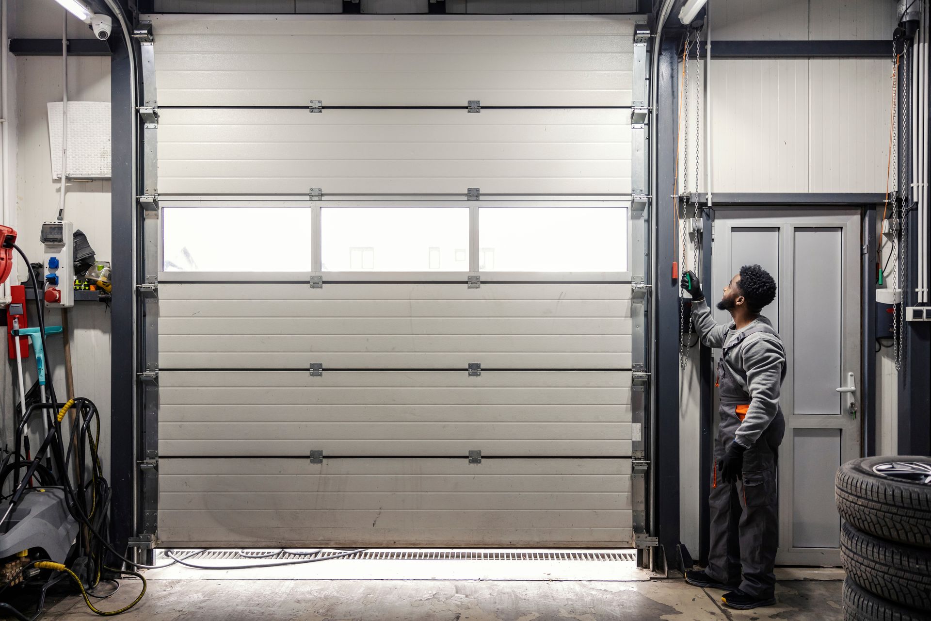 Person in a garage looking at a partially open white garage door with windows; tools and tires visible. Person in a garage looking at a partially open white garage door with windows; tools and tires visible.