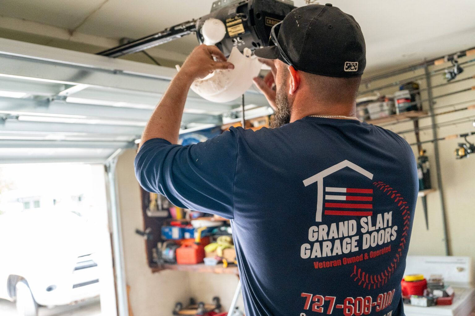 Man in blue shirt, wearing a cap, installing a garage door opener inside a garage.