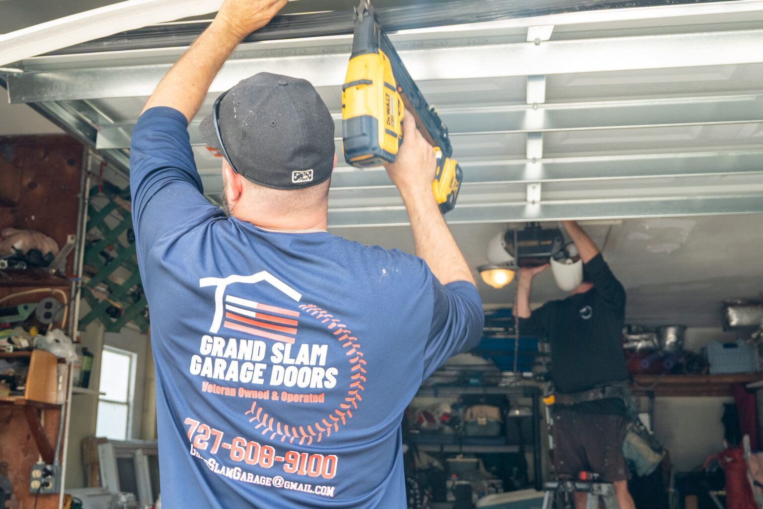 Two men installing a garage door. One uses a drill, the other works on the door's structure inside a garage.