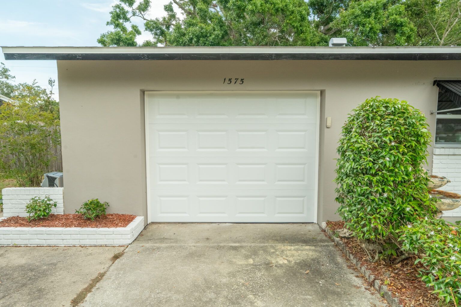 Garage of a tan-colored house with a white door. Landscaping includes small bushes and a concrete driveway.