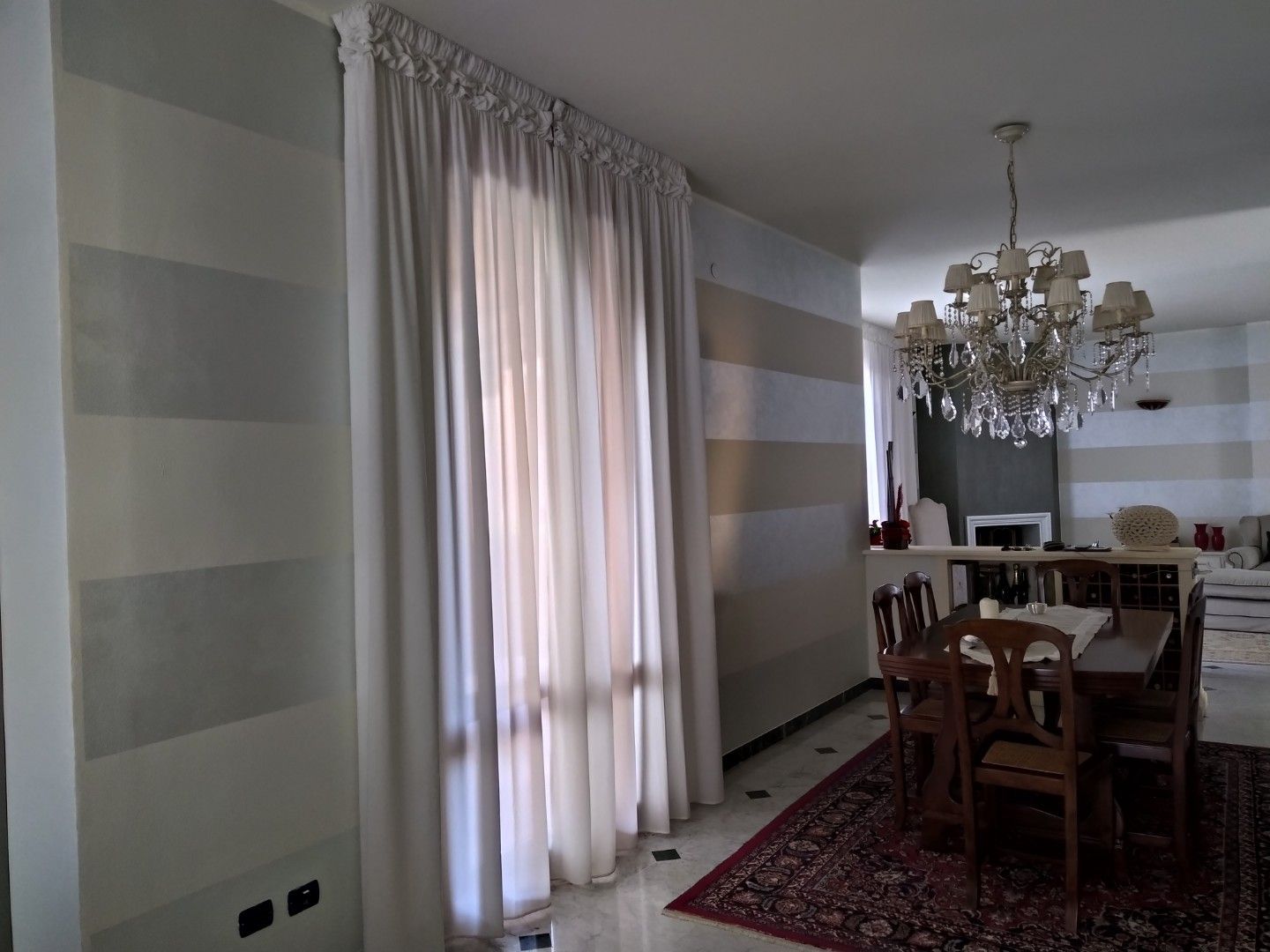 Una sala da pranzo caratterizzata da pareti con ampie strisce orizzontali beige e crema, tende fluide e un lampadario di cristallo.