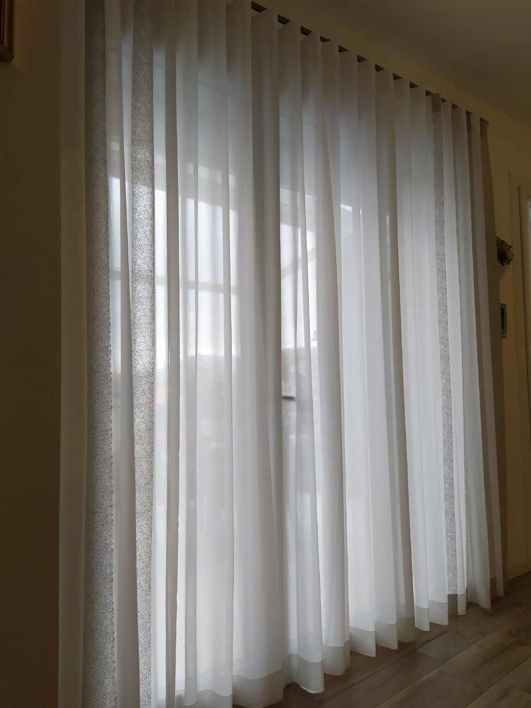 Delle tende trasparenti color avorio chiaro, con strisce verticali decorate, coprono una finestra illuminata dalla luce naturale.