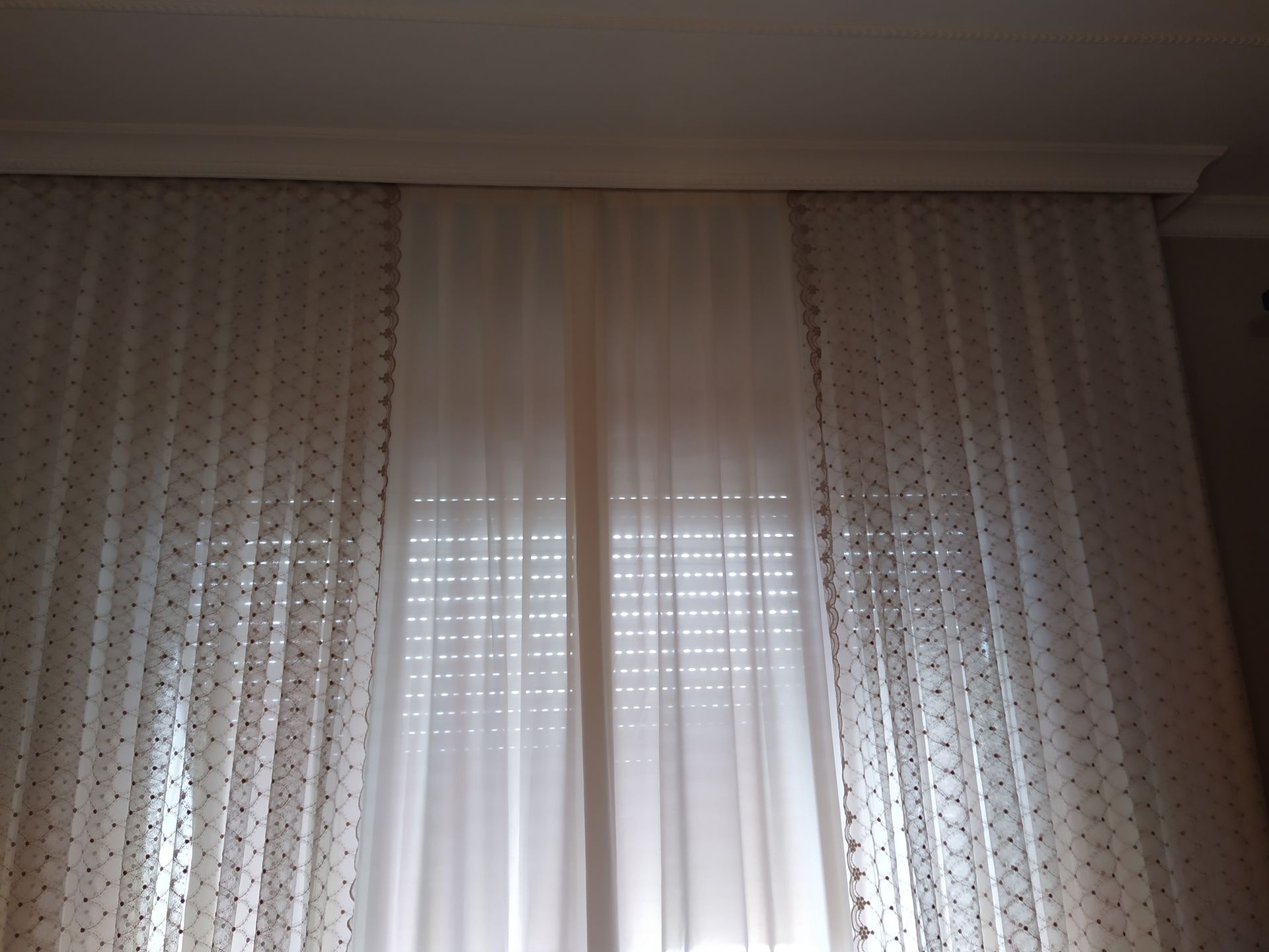 Una finestra coperta da tende decorative bianche a motivi geometrici, appese a un binario sul soffitto.