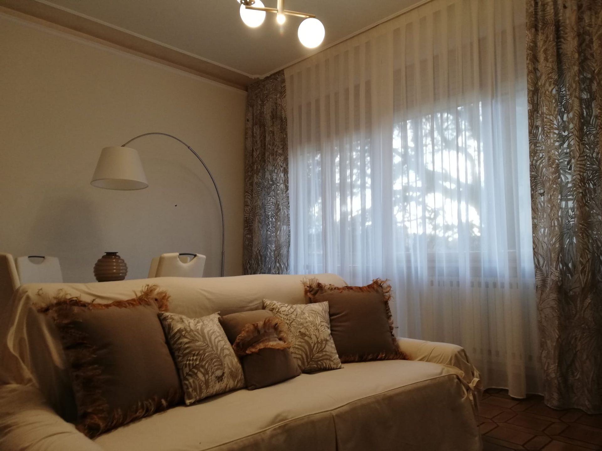 Un soggiorno con un divano chiaro dai cuscini marroni davanti a una finestra con tende trasparenti e drappeggi a fantasia.