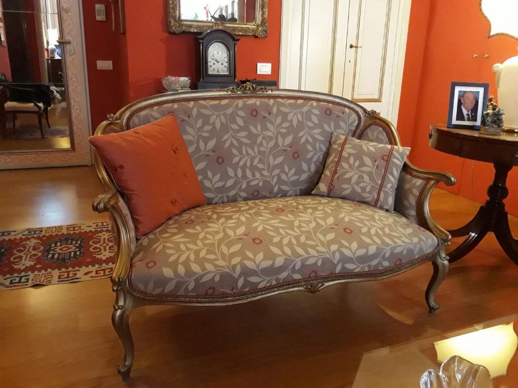 Un divanetto in stile vintage con tessuto a fantasia floreale e due cuscini decorativi in ​​una stanza con pareti arancioni e pavimento in legno.