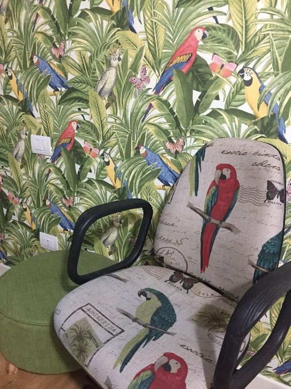 Una sedia da scrivania con un cuscino in tessuto con stampa di pappagalli è posizionata davanti a una parete ricoperta di foglie tropicali e di pappagalli dello stesso colore.