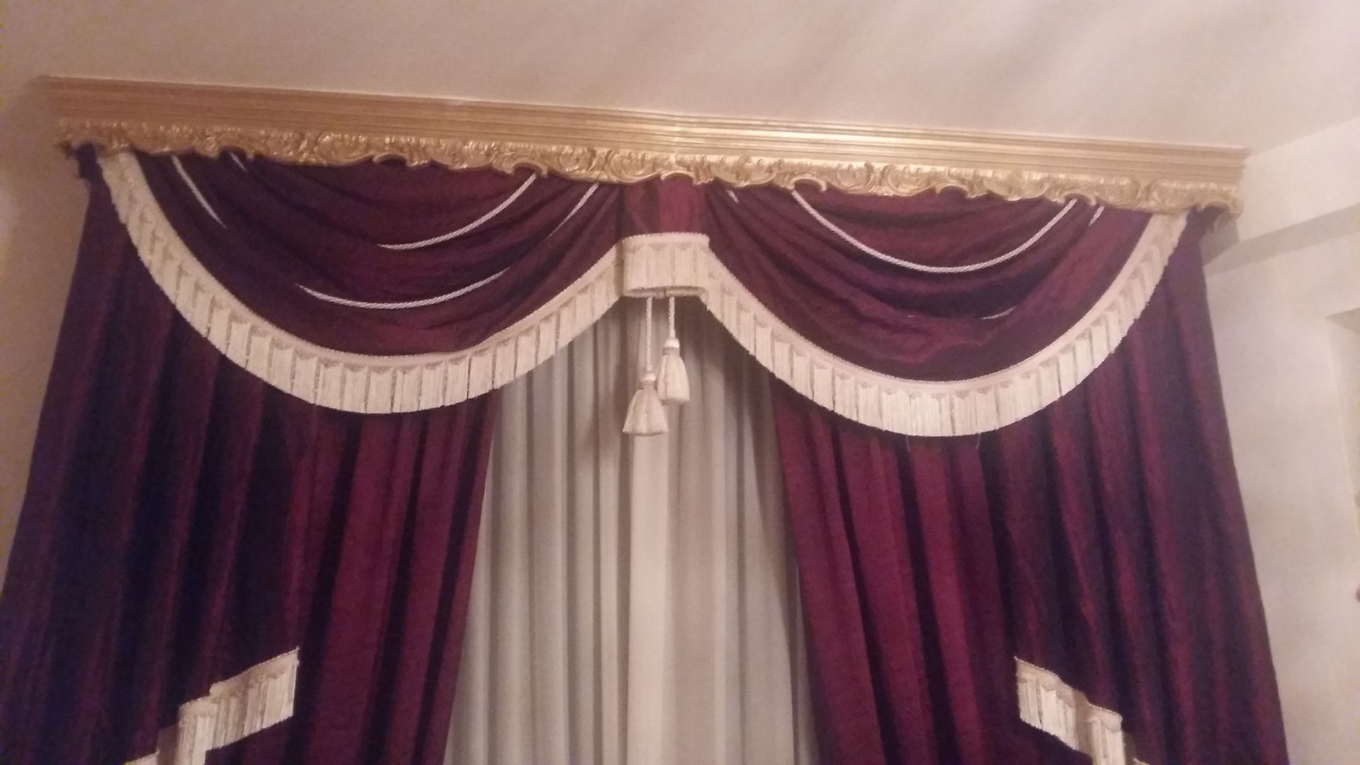 Tende color bordeaux intenso con elaborate mantovane smerlate e frange color crema, poste sotto una cornice decorativa dorata.