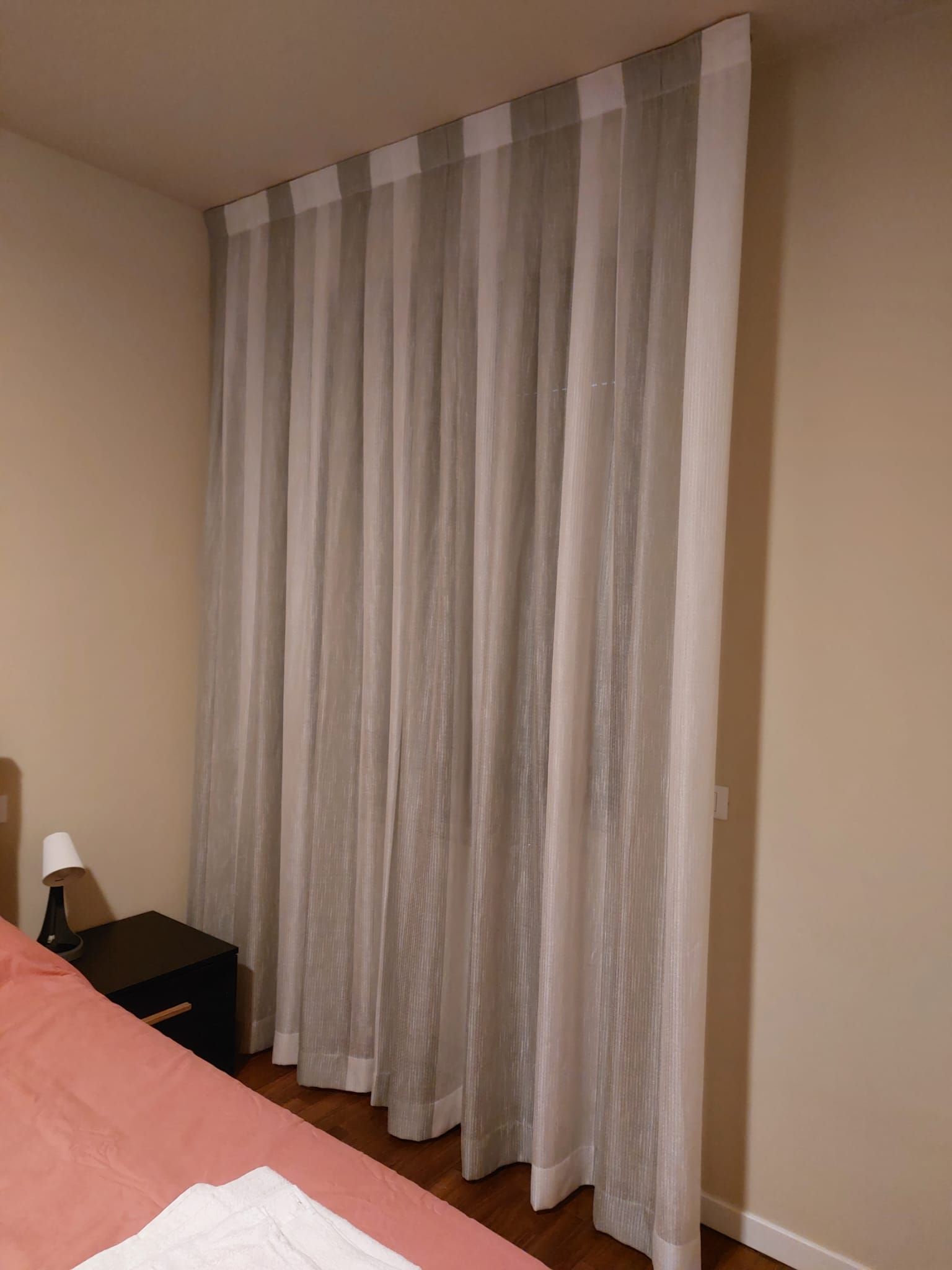 In una camera da letto, accanto al comodino e al letto, sono appese tende a righe beige e bianche che arrivano fino al soffitto.