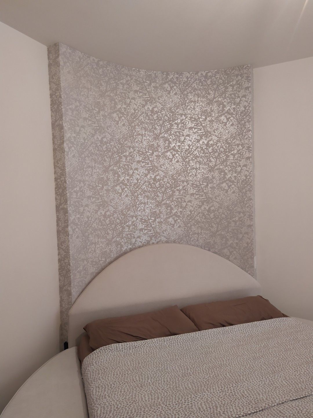 Un letto con testiera arrotondata beige, addossato a una parete caratterizzata da un pannello decorativo metallico argentato e dalla texture particolare.
