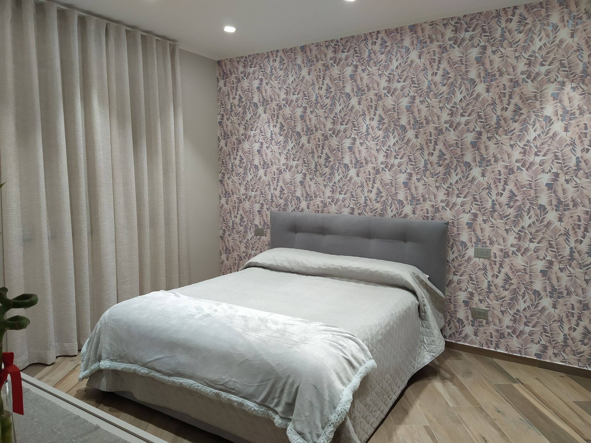 Una camera da letto con un letto grigio su una parete decorata con motivi floreali e tende chiare che arrivano fino al soffitto.