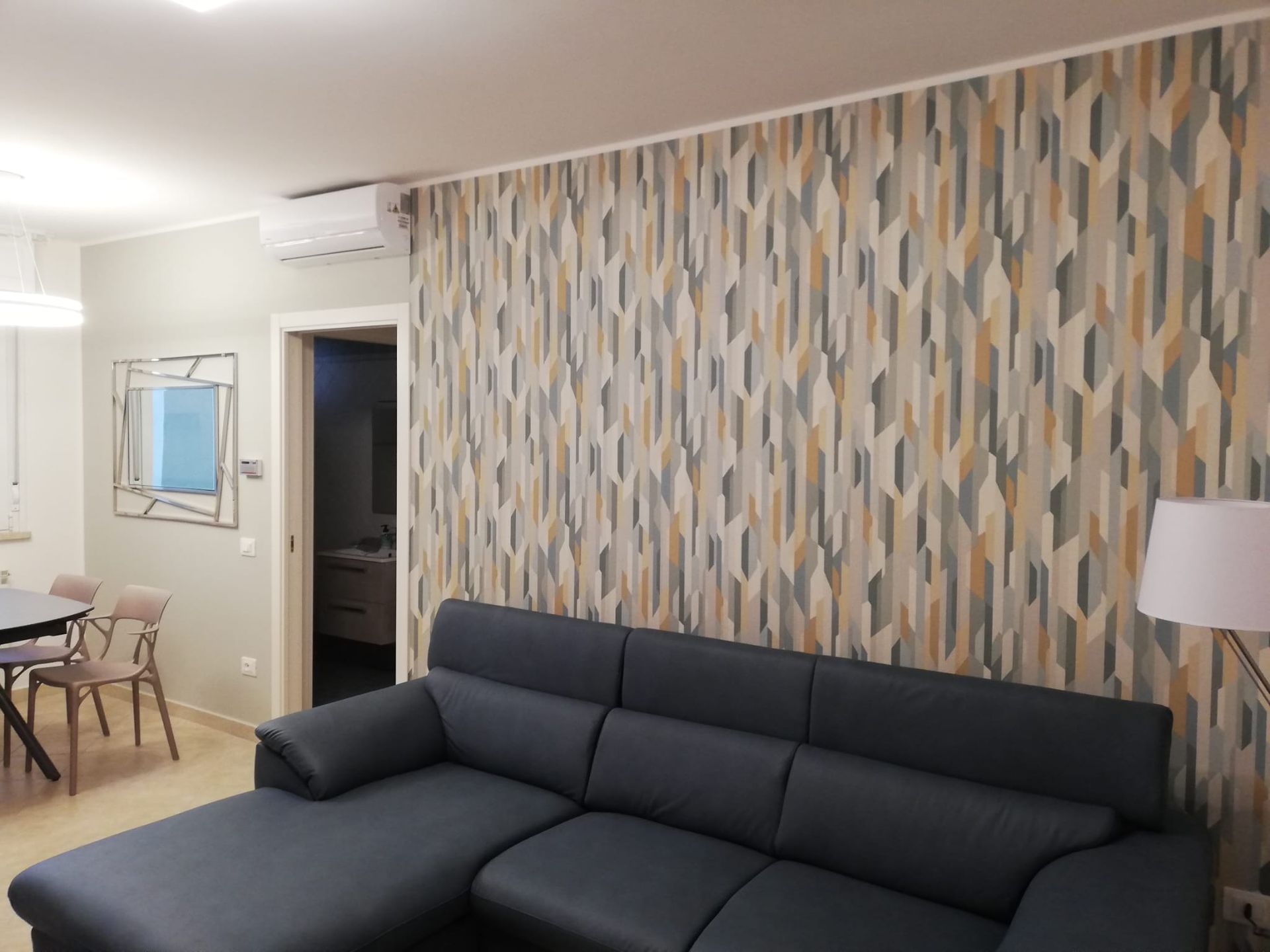 In un soggiorno, un divano componibile grigio scuro è addossato a una parete rivestita con carta da parati astratta, geometrica e a motivi verticali.