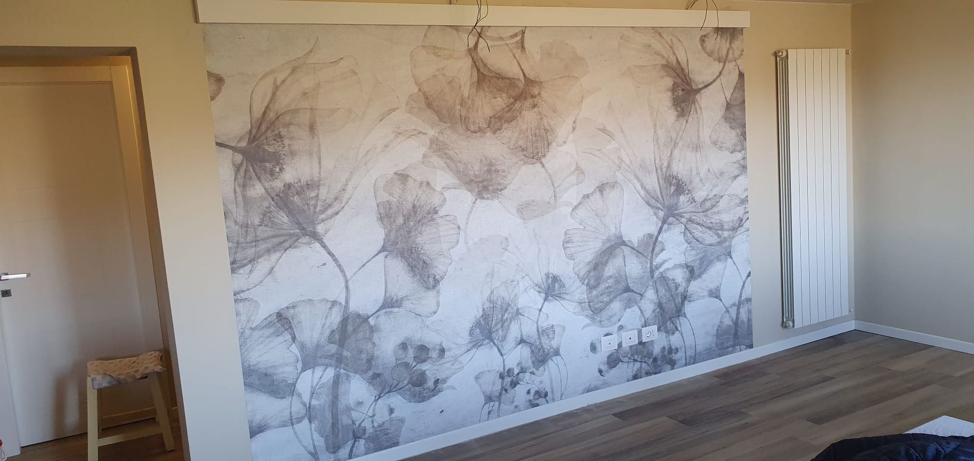 Un grande murale con motivi floreali in tonalità di grigio neutro su una parete della camera da letto, accanto a un termosifone verticale.