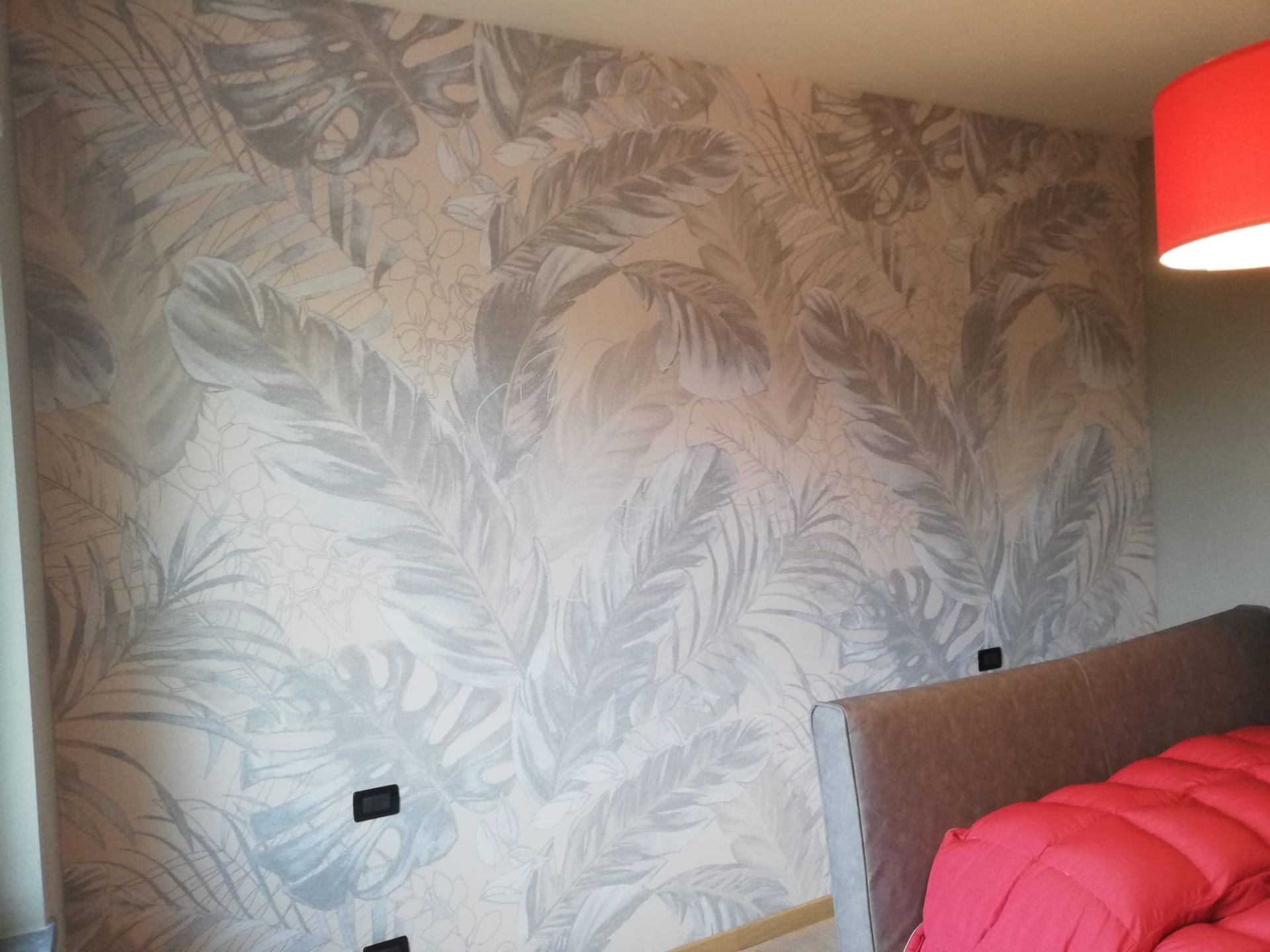 Una parete della camera da letto con carta da parati a grandi motivi di foglie di palma grigie, accanto a una testiera con lenzuola rosso acceso.